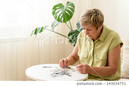 Senior woman actively engages her mind by putting together a puzzle on small table. Slow, mindful living, analog hobby, relaxing activity at home Senior woman actively engages her mind by putting together a puzzle on small table. Slow, mindful living, analog hobby, relaxing activity at home 143034751