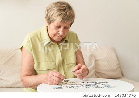 Older woman concentrating on jigsaw puzzle at white table, enjoying peaceful, brain-stimulating hobby for healthy aging Older woman concentrating on jigsaw puzzle at white table, enjoying peaceful, brain-stimulating hobby for healthy aging 143034770