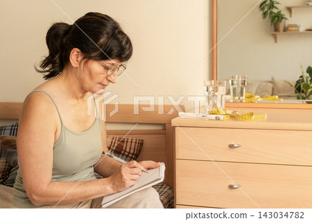 Mature woman sits at home recording data on her health and weight management progress. Consciously checking one's metrics, tracking health indicators, and making lifestyle changes Mature woman sits at home recording data on her health and weight management progress. Consciously checking one's metrics, tracking health indicators, and making lifestyle changes 143034782