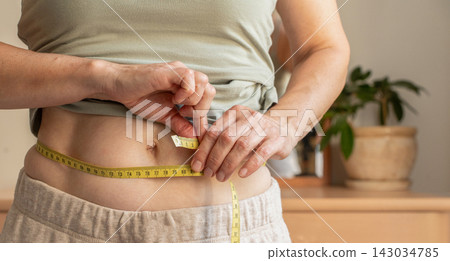 Close-up, woman measuring her waist with yellow tape measure at home. Concepts include weight loss progress, body measurements, weight management, health tracking, and lifestyle changes Close-up, woman measuring her waist with yellow tape measure at home. Concepts include weight loss progress, body measurements, weight management, health tracking, and lifestyle changes 143034785
