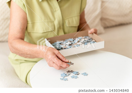An elderly womans hands select puzzle pieces from box as she enjoys quiet relaxation, cognitive exercise, and focused puzzle solving at home. Slow living, analog hobby An elderly womans hands select puzzle pieces from box as she enjoys quiet relaxation, cognitive exercise, and focused puzzle solving at home. Slow living, analog hobby 143034840