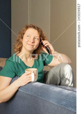 Red-haired young woman smiling while talking on phone and enjoying hot drink on sofa in morning light 143034917