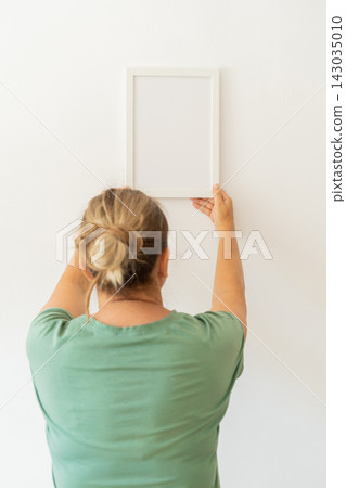 Blonde woman in green t-shirt hanging empty picture frame on white wall. Modern minimalist mockup, empty template for text Blonde woman in green t-shirt hanging empty picture frame on white wall. Modern minimalist mockup, empty template for text 143035010