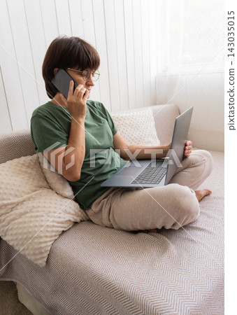 Caucasian mature female working from home on laptop and phone in cozy living room setting Caucasian mature female working from home on laptop and phone in cozy living room setting 143035015