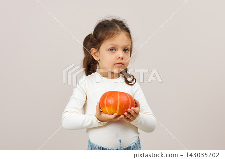 Little girl in white sweater holding an orange pumpkin. Representing childhood, autumn, and harvest concepts Little girl in white sweater holding an orange pumpkin. Representing childhood, autumn, and harvest concepts 143035202