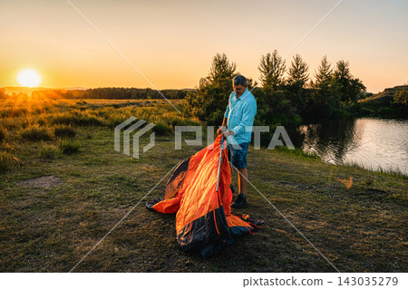 Man pitches an orange tent by calm river during golden hour, enjoying freedom, adventure, nature during his summer vacation. Intentional Solitude Man pitches an orange tent by calm river during golden hour, enjoying freedom, adventure, nature during his summer vacation. Intentional Solitude 143035279