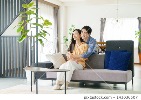 一位年輕的日本男子從背後擁抱了一位坐在客廳裡的年輕日本女子。 一位年輕的日本男子從背後擁抱了一位坐在客廳裡的年輕日本女子。 143036507