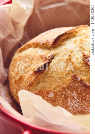 Golden sourdough bread with cracked crust in red ceramic Dutch oven, lined with parchment paper. Perfect for culinary, baking, and food photography projects. Golden sourdough bread with cracked crust in red ceramic Dutch oven, lined with parchment paper. Perfect for culinary, baking, and food photography projects. 143038305