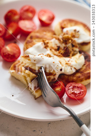 Savory syrniki (cheese pancakes) topped with sour cream, crispy fried onions, and fresh cherry tomatoes, served on a white plate with a blue knit cloth. Perfect for breakfast or brunch. Savory syrniki (cheese pancakes) topped with sour cream, crispy fried onions, and fresh cherry tomatoes, served on a white plate with a blue knit cloth. Perfect for breakfast or brunch. 143038313