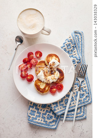 Savory syrniki (cheese pancakes) topped with sour cream, crispy fried onions, and fresh cherry tomatoes, served on a white plate with a blue knit cloth. Perfect for breakfast or brunch. Savory syrniki (cheese pancakes) topped with sour cream, crispy fried onions, and fresh cherry tomatoes, served on a white plate with a blue knit cloth. Perfect for breakfast or brunch. 143038329