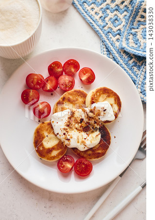 Savory syrniki (cheese pancakes) topped with sour cream, crispy fried onions, and fresh cherry tomatoes, served on a white plate with a blue knit cloth. Perfect for breakfast or brunch. Savory syrniki (cheese pancakes) topped with sour cream, crispy fried onions, and fresh cherry tomatoes, served on a white plate with a blue knit cloth. Perfect for breakfast or brunch. 143038330