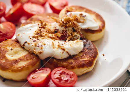 Savory syrniki (cheese pancakes) topped with sour cream, crispy fried onions, and fresh cherry tomatoes, served on a white plate with a blue knit cloth. Perfect for breakfast or brunch. 143038331