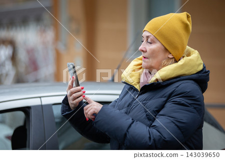 Woman wearing yellow hat checks smartphone outside her car in a city during winter Woman wearing yellow hat checks smartphone outside her car in a city during winter 143039650