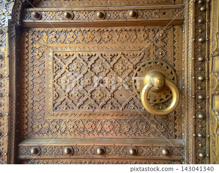 The fragment depicts a gilded wooden door decorated with fine carvings and a bronze ring. Traditional Indian decorative art, Jaipur. The fragment depicts a gilded wooden door decorated with fine carvings and a bronze ring. Traditional Indian decorative art, Jaipur. 143041340