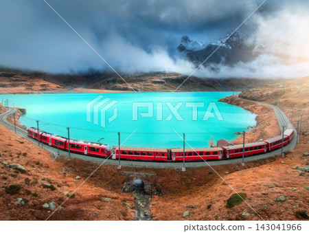 Aerial view of red train near alpine mountain lake and cloudy sky Aerial view of red train near alpine mountain lake and cloudy sky 143041966