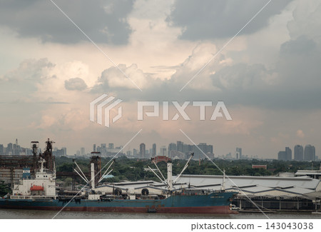 Moored along the Chao Phraya River, a large industrial vessel sits framed by the lush foliage of Bang Kachao, distant city skyscrapers, and a dramatic cloudscape. Moored along the Chao Phraya River, a large industrial vessel sits framed by the lush foliage of Bang Kachao, distant city skyscrapers, and a dramatic cloudscape. 143043038