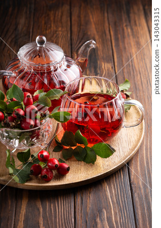 Glass teapot and cup filled with rosehip tea alongside fresh berries and leaves on a wooden tray Glass teapot and cup filled with rosehip tea alongside fresh berries and leaves on a wooden tray 143043315
