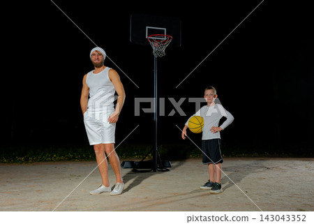 Smiling adult parent with cute child coming at outdoor court to play basketball. Father and son with ball talking while walking. Playground in city park. Smiling adult parent with cute child coming at outdoor court to play basketball. Father and son with ball talking while walking. Playground in city park. 143043352