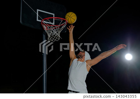 A man performs a slam dunk on a basketball court at night under the glare of bright outdoor lights A man performs a slam dunk on a basketball court at night under the glare of bright outdoor lights 143043355