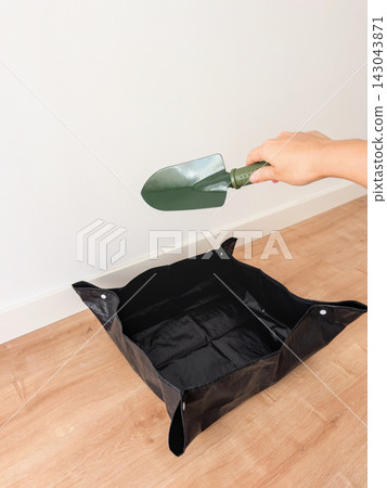 Woman's hand holding a green garden trowel above an empty black repotting mat on a wooden floor Woman's hand holding a green garden trowel above an empty black repotting mat on a wooden floor 143043871