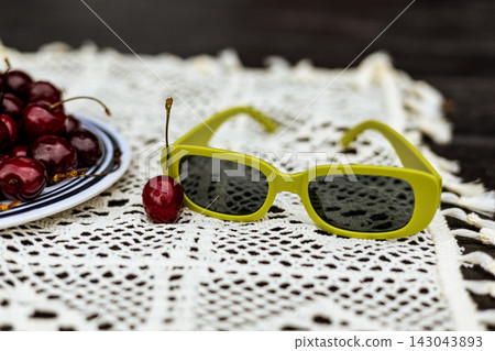 National Picnic Month. Trendy yellow green sunglasses on a white lace tablecloth with cherries. Retro summer fashion, cottagecore, vintage vacation. National Picnic Month. Trendy yellow green sunglasses on a white lace tablecloth with cherries. Retro summer fashion, cottagecore, vintage vacation. 143043893
