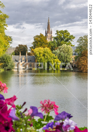 Bruges town in Belgium, Europe. Historic European cityscape with prominent architectural spire behind a tranquil lake, framed by vibrant flowers and lush autumn trees 143044622