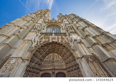 Antwerp town in Belgium, Europe. Cathedral of Our Lady in Antwerp, low angle view of an ancient towering cathedral facade against a vibrant blue sky with wispy clouds, showcasing gothic architecture Antwerp town in Belgium, Europe. Cathedral of Our Lady in Antwerp, low angle view of an ancient towering cathedral facade against a vibrant blue sky with wispy clouds, showcasing gothic architecture 143044624
