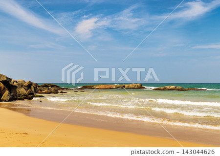 Idyllic coastal scene with golden sand beach, clear blue sky, and rocks along the tranquil turquoise ocean shore on a sunny day. Sri Lanka island in Asia. Idyllic coastal scene with golden sand beach, clear blue sky, and rocks along the tranquil turquoise ocean shore on a sunny day. Sri Lanka island in Asia. 143044626