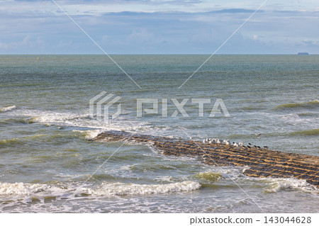 Panoramic view of a textured rock ridge extending into the vast, choppy sea under a dynamic sky with scattered clouds and distant horizon. Ostend town in Belgium, Europe. Panoramic view of a textured rock ridge extending into the vast, choppy sea under a dynamic sky with scattered clouds and distant horizon. Ostend town in Belgium, Europe. 143044628