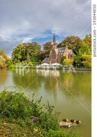 Bruges town in Belgium, Europe. Minnewater Castle, Historic building with spire by a serene lake, surrounded by vibrant autumn trees under a soft, cloudy sky, evoking peace Bruges town in Belgium, Europe. Minnewater Castle, Historic building with spire by a serene lake, surrounded by vibrant autumn trees under a soft, cloudy sky, evoking peace 143044650