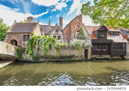 Bruges town in Belgium, Europe. Historic European architectural scenery with ancient buildings, stone bridge, and canal under a vibrant blue sky with clouds Bruges town in Belgium, Europe. Historic European architectural scenery with ancient buildings, stone bridge, and canal under a vibrant blue sky with clouds 143044659
