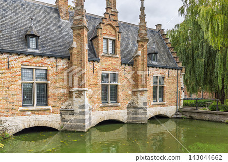 Bruges town in Belgium, Europe. Historic brick structure with arched foundations over tranquil green canal water, beside a weeping tree, under a soft sky Bruges town in Belgium, Europe. Historic brick structure with arched foundations over tranquil green canal water, beside a weeping tree, under a soft sky 143044662