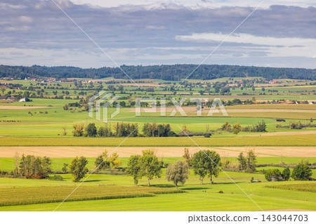 Expansive Rural Landscape with Green Fields, Distant Forest, and Scattered Settlements Under a Cloudy Sky. Region of Bavaria in Germany, Europe. Expansive Rural Landscape with Green Fields, Distant Forest, and Scattered Settlements Under a Cloudy Sky. Region of Bavaria in Germany, Europe. 143044703