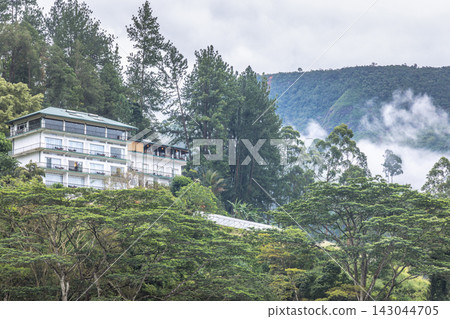 Modern hotel architecture nestled amidst green mountain forest with mist and cloudy sky on a serene day. Mountain landscape near Nuwara Eliya in Sri Lanka. Modern hotel architecture nestled amidst green mountain forest with mist and cloudy sky on a serene day. Mountain landscape near Nuwara Eliya in Sri Lanka. 143044705