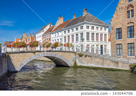Bruges town in Belgium, Europe. Charming canal scene with historic architecture, stone bridge adorned with flower baskets, and vibrant blue sky on a sunny day Bruges town in Belgium, Europe. Charming canal scene with historic architecture, stone bridge adorned with flower baskets, and vibrant blue sky on a sunny day 143044706