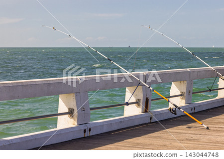 Two fishing rods with reels rest on a wooden pier overlooking a vast blue sea under a bright, clear sky, inviting relaxation. Nieuwpoort town in Belgium, Europe. Two fishing rods with reels rest on a wooden pier overlooking a vast blue sea under a bright, clear sky, inviting relaxation. Nieuwpoort town in Belgium, Europe. 143044748