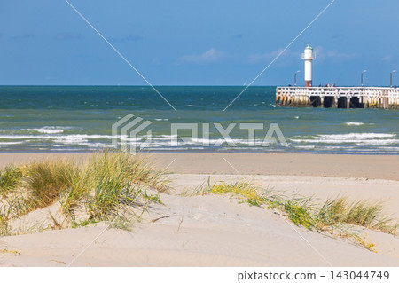 Sunny Coastal Landscape with Sandy Beach, Green Dunes, and Distant Lighthouse on a Pier by the Blue Sea under Clear Sky. Nieuwpoort town in Belgium, Europe. Sunny Coastal Landscape with Sandy Beach, Green Dunes, and Distant Lighthouse on a Pier by the Blue Sea under Clear Sky. Nieuwpoort town in Belgium, Europe. 143044749