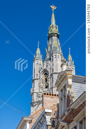 Ghent town in Belgium, Europe. Stone Bell Tower with Ornate Spire and Golden Weather Vane Soars Against a Bright Blue Sky, Overlooking Historic Rooftops Ghent town in Belgium, Europe. Stone Bell Tower with Ornate Spire and Golden Weather Vane Soars Against a Bright Blue Sky, Overlooking Historic Rooftops 143044768