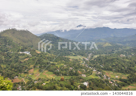 Panoramic view of lush green mountain landscape with distant peaks shrouded in clouds under a dramatic sky. Mountain landscape near Ella town in Sri Lanka island in Asia. Panoramic view of lush green mountain landscape with distant peaks shrouded in clouds under a dramatic sky. Mountain landscape near Ella town in Sri Lanka island in Asia. 143044816