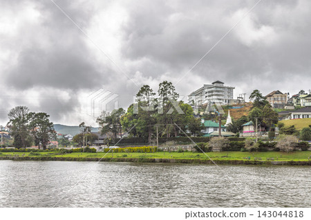 Serene Lakeside View of a Hillside Town with Lush Greenery and Overcast Skies, Reflecting Tranquility and Urban Nature Harmony. Nuwara Eliya town in Sri Lanka. Serene Lakeside View of a Hillside Town with Lush Greenery and Overcast Skies, Reflecting Tranquility and Urban Nature Harmony. Nuwara Eliya town in Sri Lanka. 143044818