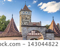 Rothenburg ob der Tauber, medieval old town in region of Bavaria in Germany, Europe. Historic Stone Archway and Medieval Tower with Terracotta Roofs Against Blue Sky 143044822