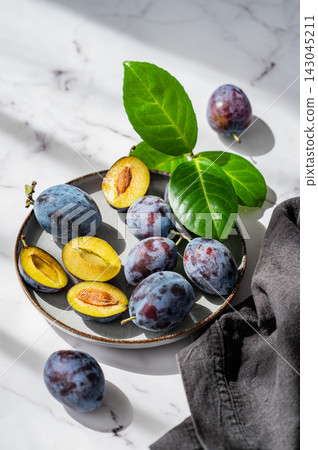 Fresh blue plum whole and sliced on a plate with green leaves on marble background with sunlight. Fresh blue plum whole and sliced on a plate with green leaves on marble background with sunlight. 143045211