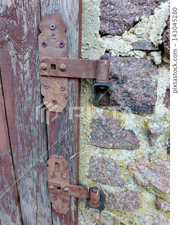 An old, rusty metal hinge on the wooden door of a stone building. An old, rusty metal hinge on the wooden door of a stone building. 143045280