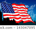 An American flag flutters against a backdrop of a blue sky and the silhouette of a city. 143047095