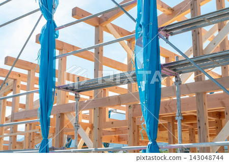 A new wooden house under construction, surrounded by scaffolding, is being built. The columns and beams of the custom-built house are being assembled. A new wooden house under construction, surrounded by scaffolding, is being built. The columns and beams of the custom-built house are being assembled. 143048744