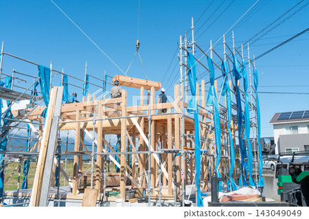 The framing of a newly built wooden house. A carpenter lifts a beam with a crane, and construction work is underway. The framing of a newly built wooden house. A carpenter lifts a beam with a crane, and construction work is underway. 143049049