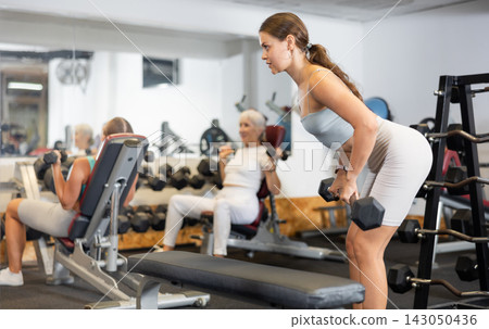Slender girl trains muscles of arms and back using barbell and differentiated weighting in gym Slender girl trains muscles of arms and back using barbell and differentiated weighting in gym 143050436