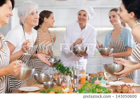 Aged woman holding wine and talking to other cooking course members whipping souce in bowls Aged woman holding wine and talking to other cooking course members whipping souce in bowls 143050600
