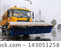 Yellow snowplow truck equipped with a wide blue brush clearing snow and slush from a city street during winter 143051258