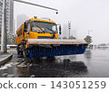 Yellow snowplow truck equipped with a wide blue brush clearing snow and slush from a city street during winter 143051259